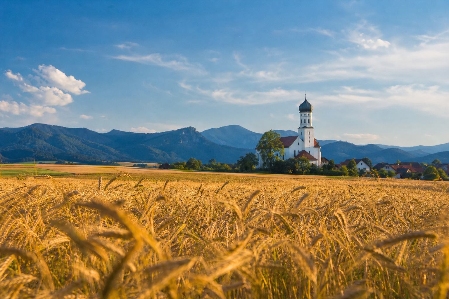 Goldene Felder und bayerisches Dorf mit Holzhäusern vor blauen Hügeln
